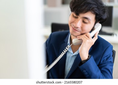 Asian Male Financial Advisor, Operator Or Telemarketer Talking On The Phone With Customer And Working With Computer At Desk In The Office. 