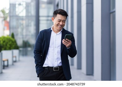 asian male freelancer walking near business center holding phone, smiling reading news, successful businessman - Shutterstock ID 2048743739