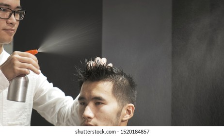 Asian Male Is Getting A Modern Haircut In Barber Shop. Barber Wets Hair By Spray And Combs Them.