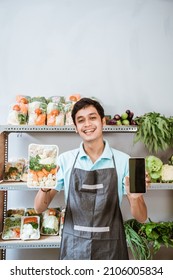 Asian Male Greengrocer Showing Vegetables And Showing A Phone Screen