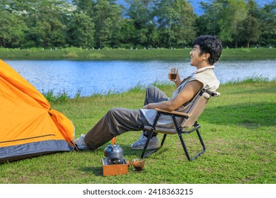 Asian male man sitting on chair, drinking coffee and feeling relax with camping tent beside lake and mountain. - Powered by Shutterstock