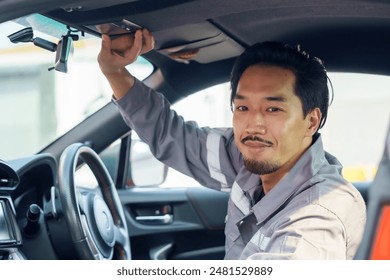 Asian male mechanic adjusting rearview mirror inside a car. Wearing a grey uniform. Represents vehicle inspection, professional service, and automotive maintenance in a workshop environment