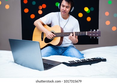 Asian Male Musician Composing A Song With Laptop Computer & Midi Keyboard, Practicing Guitar With Online Music Lesson On White Bed At Night. Song Writing, Music Education & Bedroom Studio Concept