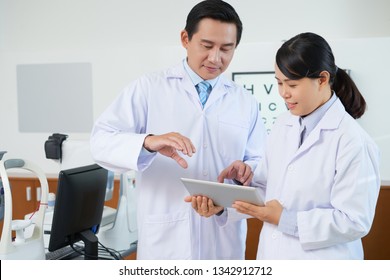 Asian male ophthalmologist using digital tablet together with his assistant, he standing and explaining to her the new technologies in optometry - Powered by Shutterstock
