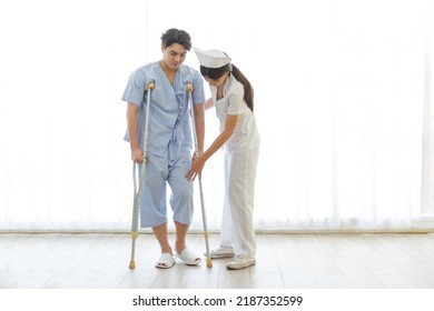 Asian Male Patient Walking Slowly With Crutch While Nurse Supporting At Hospital Ward. Caregiver Therapist Nurse Support  Male Use Crutch. Medical Therapy Insurance Concept.