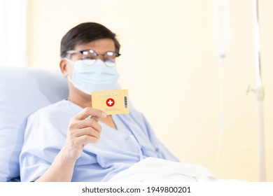 Asian Male Patients Wearing Protective Hygienic Mask And Showing Health Card For Medical Expenses On A Hospital Bed. Elderly Health Insurance, Life Insurance And Financial Planning