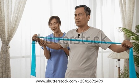 asian male pensioner exercising with resistance tape and the support of the woman caregiver at home. he pulls it with all his exertion at daytime