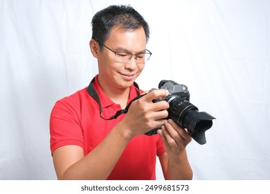 An Asian male photographer wearing red shirt holding camera and review the photos on the camera screen with white background. Travel concept - Powered by Shutterstock