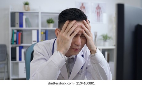 Asian Male Physician Under Pressure At Work Is Burying His Face With Hands And Breathing A Deep Sigh While Looking At The Computer Monitor With Anxiety.