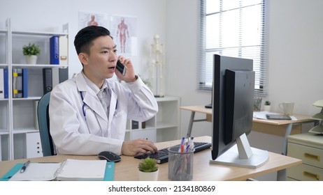 Asian Male Physician Working In Front Of The Computer Is Talking In Surprise And Leaving His Office Right Away As Soon As He Receives An Emergency Call.