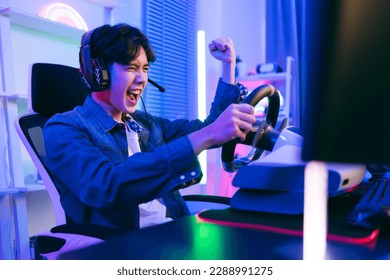 Asian male pro gamer playing an online car drive simulation game with steering wheel controller. Gamer winning an sports game with victory emotion. The player rejoices in victory in the competition. - Shutterstock ID 2288991275
