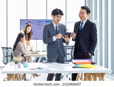 Asian male professional businessmen colleague in formal business suit take coffee break standing holding disposable paper cup and tablet computer talking have conversation together in front monitor.