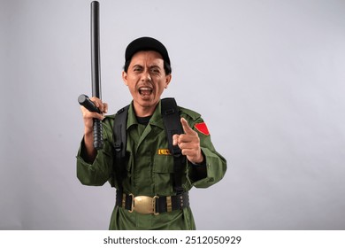 Asian male security officer in a green uniform holding a baton and pointing with a firm, assertive expression. He is wearing tactical gear and a black cap, standing against a plain background. - Powered by Shutterstock