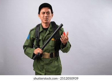 Asian male security officer in a green uniform holding a baton, making a confused or questioning expression. He is equipped with tactical gear, wearing a black cap, against a plain background. - Powered by Shutterstock