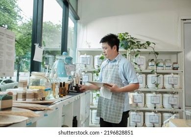 Asian Male Shopkeeper Checks Stock Of Natural Organic Products At Window Display In Refill Store, Zero-waste And Plastic-free Grocery, Eco Environment-friendly, Sustainable Lifestyles, Reusable Shop.