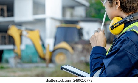 Asian male site engineer or building inspector man with green reflective vest, safety goggles and earmuffs thinking about working process while using digital tablet at construction site. - Powered by Shutterstock