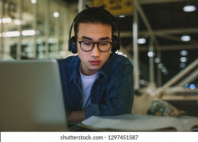 Asian Male Student With Headphones Sitting At Library Reading Book And Using Laptop. Man Studying At University Library.