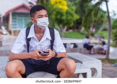 An Asian Male Student In White High School Uniforms Wearing The Mask Use Mobile Phone While Waiting For Car To Go Back Home After School During The Coronavirus 2019 (Covid-19) Epidemic.