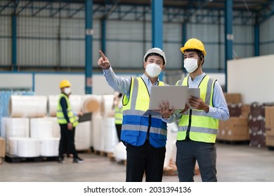 Asian Male Supervisor Engineer In Helmet Safety Wearing Hygienic Mask To Protect Coronavirus With Technician Holding Laptop Point To Storage Location Of Products In Warehouse Factory Industrial.