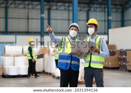 Asian male supervisor engineer in helmet safety wearing hygienic mask to protect coronavirus with technician holding laptop point to storage location of products in warehouse factory industrial.