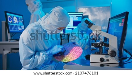 asian male technician in clean room suit holds wafer that reflects many different colors with gloves and check it at semiconductor manufacturing plant