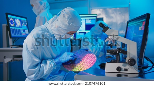 asian male technician in clean room\
suit holds wafer that reflects many different colors with gloves\
and check it at semiconductor manufacturing\
plant