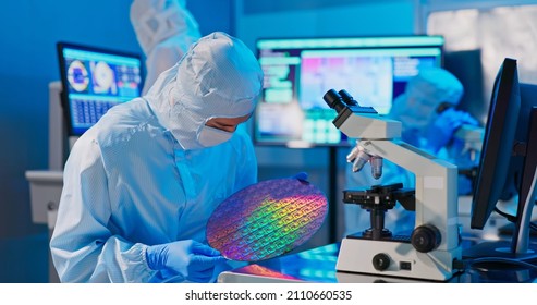asian male technician in sterile coverall holds wafer that reflects many different colors with gloves and check it at semiconductor manufacturing plant - Shutterstock ID 2110660535