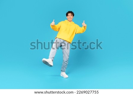 Asian Male Teen Gesturing Thumbs Up With Both Hands Posing On Blue Studio Background. Teenager Approving Great Offer With Like Gestures Smiling To Camera. Full Length Shot