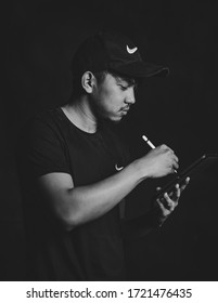 Asian Male using iPad tablet and pencil to edit photo. Man wearing cap.