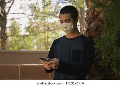 Asian Male Wearing Mask Standing Outside Surrounded By Trees Looking Down At His Cell Phone