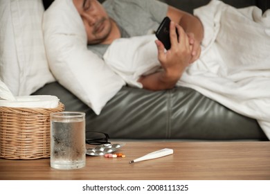 Asian Man Is Absent From Work And Sick Leave Because Of Illness, Takes Temperature By Thermometer, Takes Medicine, And Sleeps In Blankets On Sofa At Home, Tissues, And Capsule Pills On The Table.