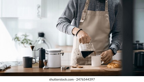 Asian man in apron pouring coffee in cup while standing near cooking surface in kitchen - Powered by Shutterstock