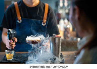 Asian Man Barista Shaking Iced Black Coffee In Shaker And Pouring In Cocktail Glass On Bar Counter At Coffee Shop. Male Coffee Shop Owner Serving Cold Drink Coffee To Customer. Small Business Concept.