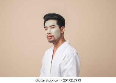 Asian Man In Bathrobe Facial Treatment And Skin Rejuvenation With Clay Mask On Face In Studio Brown Color Background.