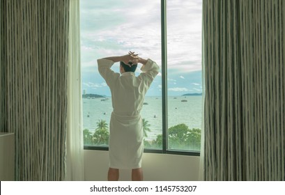 Asian Man In Bathrobe Suit Open Curtain To See View Outside In Morning, Travel And Holiday Concept