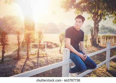 An asian man in black T-shirt and jeans sitting on the white fence and looking at the camera with sun flare effect.
