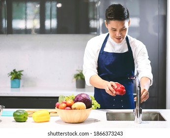 Asian man in blue apron washing red sweet pepper in the kitchen sink. - Powered by Shutterstock