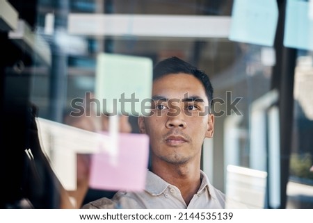 An Asian man brainstorms with sticky notes as seen through a window