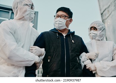 Asian Man Breaking Covid Regulation Getting Taken Away Or Arrested By Healthcare Workers Wearing Hazmat Protective Suits. Male Removed For Not Following The Rules Or Restrictions Of The Pandemic.