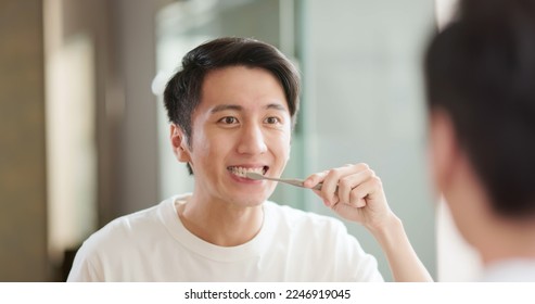 Asian man brushing teeth in bathroom to mirror