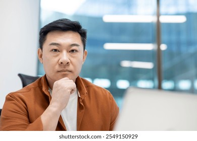 Asian man businessman seated in office setting with thoughtful expression, symbolizing decision-making, confidence, strategic thinking. Casual attire reflects modern professional environment. - Powered by Shutterstock