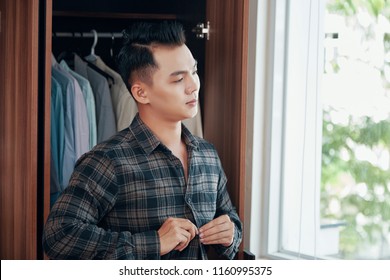 Asian Man Buttoning Checkered Shirt While Getting Dressed In Room With Wardrobe At Home Looking Away