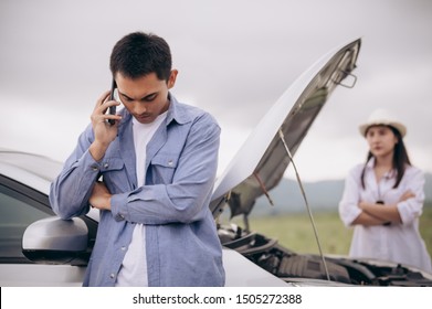 Asian man calling car maintenance service with his girlfriend looking at him background between long road trip. Car broken in mountain meadow with wife. People and transportation concept. Car repair