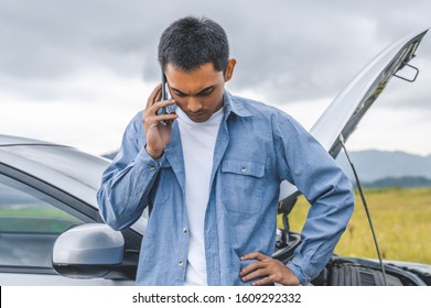 Asian Man Calling Car Maintenance Service After Having Accident During Travel Long Weekend Between Road Trip. Car Broken In Mountain Meadow Background. People Transportation. Need Help Insurance Claim