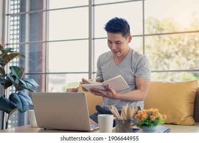 Asian Man In Casual Outfit Sitting In Living Room Reading Or Lecture Book And Using Laptop In Happy And Smile Emotion