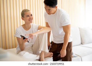 Asian Man And Caucasian Woman Sitting With Phone At Home. Concept Of Relationship And Spending Time Together. Idea Of Domestic Lifestyle. Smiling Multiracial Couple In Studio Apartment