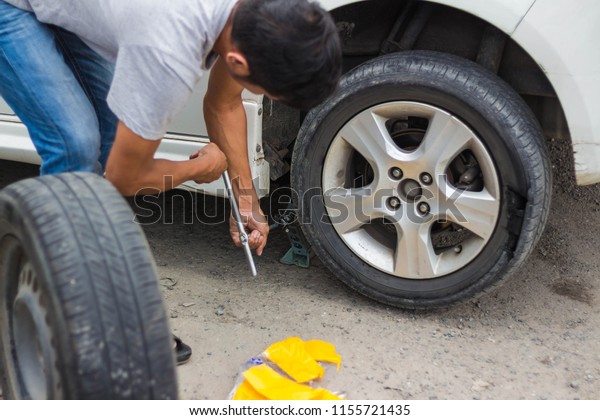 An Asian man is changing the tire. While the tire\
broke By using tools And equipment attached to the car Used to\
change the spare tire alone
