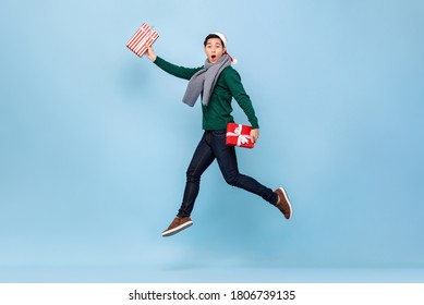 Asian Man In Christmas Sweater Jumping And Holding Gift Boxes Isolated In Light Background