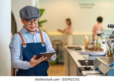 Asian Man Coffee Shop Manager Walking Behind Bar Counter Working On Digital Tablet Checking And Counting Inventory For Ordering Product For Cafe. Small Business Restaurant Owner Entrepreneur Concept