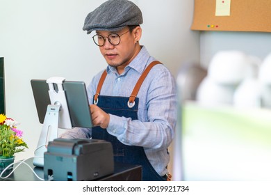 Asian Man Coffee Shop Manager Walking Behind Bar Counter Working On Digital Tablet Checking And Counting Inventory For Ordering Product For Cafe. Small Business Restaurant Owner Entrepreneur Concept